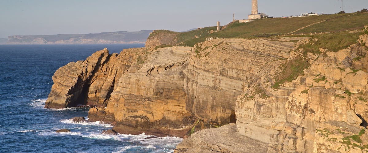 A coastal cliff with a lighthouse and rocky waves crashing against it.