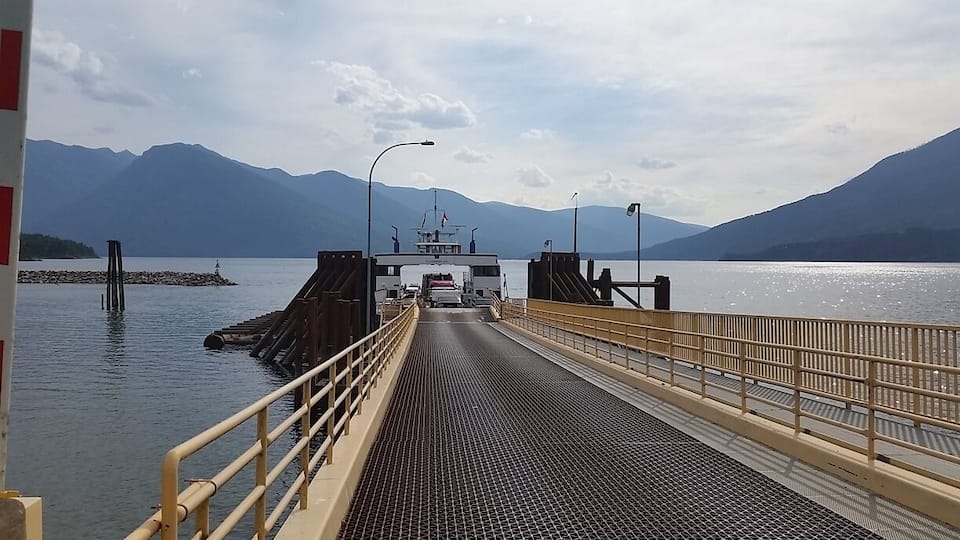 A ferry dock with a metal ramp leading to a boat, surrounded by calm water and mountains in the background.