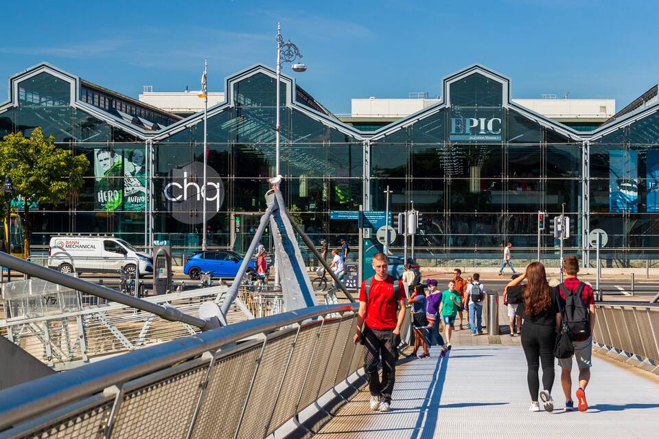 Un pont piétonnier avec, en arrière-plan, un bâtiment en verre sur lequel on peut lire les inscriptions « PIC » et « JEANIE JOHNSTON ».