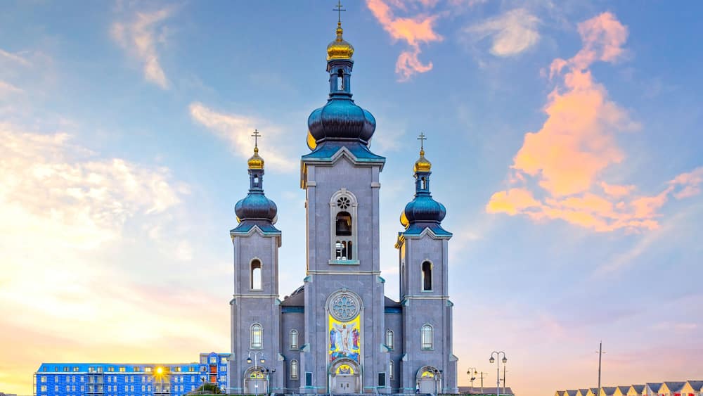 A large church with three domes and a central bell tower, surrounded by a grassy area and a row of colorful buildings.