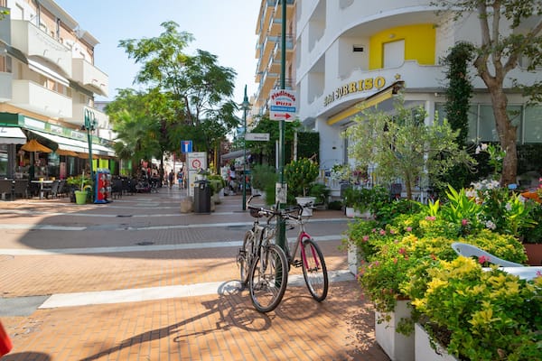 Eine Straße mit einem Fahrrad, das neben einem Blumenbeet steht, einem Schild für 'Piadiniera' und 'Pizza' sowie einem Gebäude mit der Aufschrift 'Sorriso'.