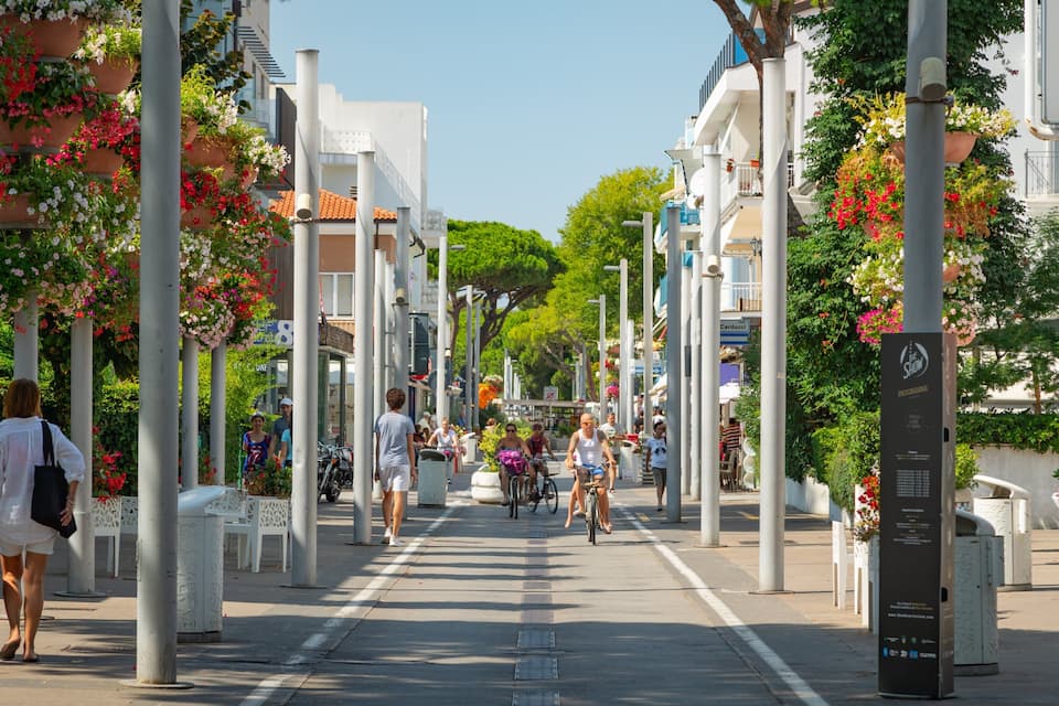A pedestrian street lined with white buildings, potted plants, and people walking and cycling.