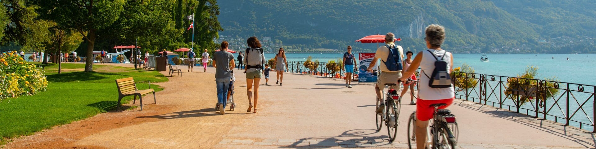 A scenic lakeside path with cyclists and pedestrians, surrounded by lush greenery and mountains.