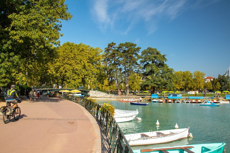 A riverside park with boats docked, a cyclist, and a bridge.