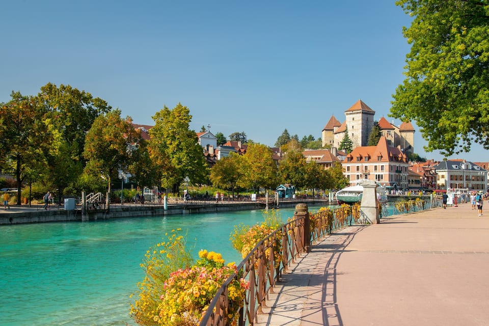 Un paseo fluvial con un río turquesa cristalino, un puente con barandilla decorada con flores y edificios históricos al fondo.