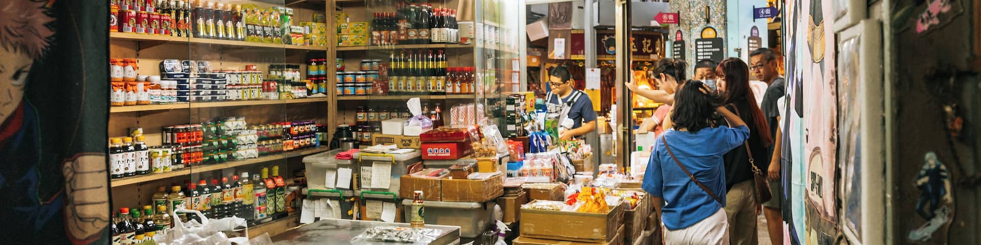 A narrow market aisle with shelves stocked with various packaged goods, a refrigerated display case, and a person standing at the counter.
