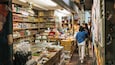Un pasillo estrecho en un mercado con estantes llenos de productos envasados, un mostrador refrigerado y una persona de pie en la barra.