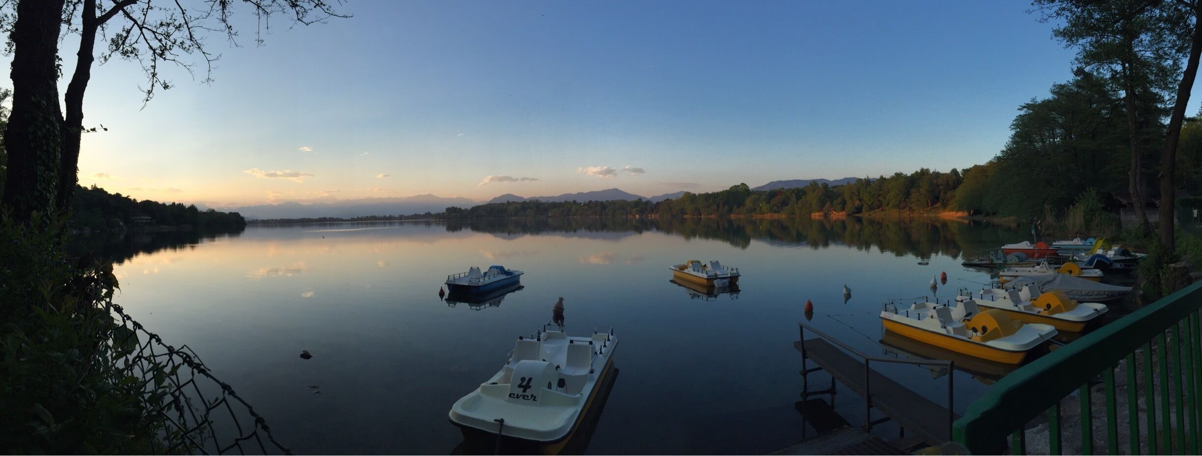 A calm lake with boats docked along the shore, surrounded by trees and mountains in the background.