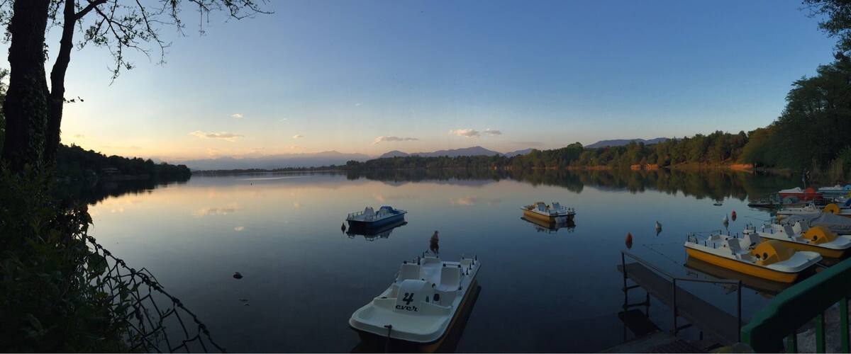 A calm lake with boats docked along the shore, surrounded by trees and mountains in the background.