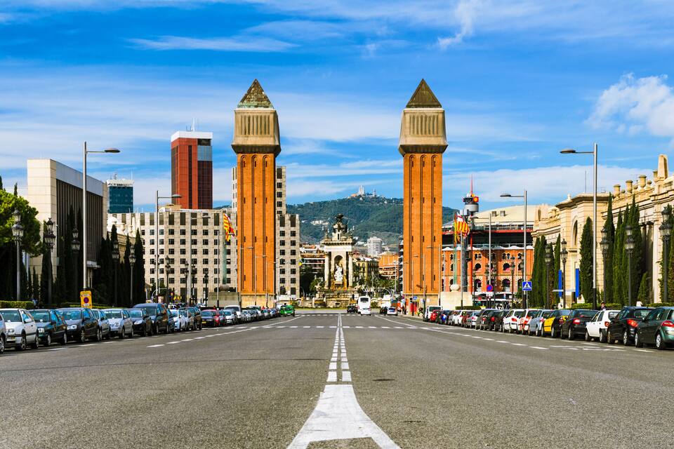 A wide city street with parked cars, tall buildings, and two prominent towers.
