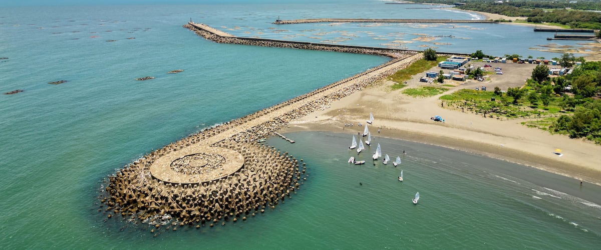 A coastal area with a circular stone structure in the water, a sandy beach, and sailboats.