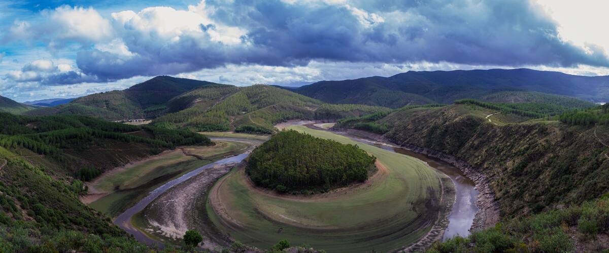 A river winding through a valley with forested hills and a cloudy sky.