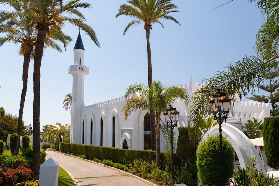 Eine Moschee mit einem hohen Minarett, umgeben von Palmen und gepflegten Gärten.