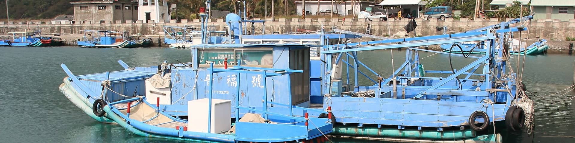 Un puerto con varias embarcaciones pesqueras azules amarradas, un faro al fondo y montañas a lo lejos.