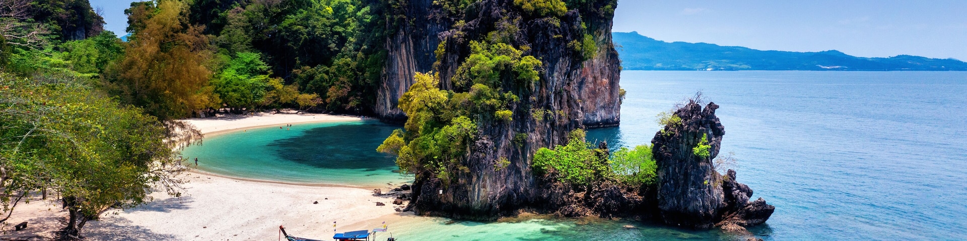 Une plage tropicale aux eaux turquoise et limpides, une grande formation rocheuse et un bateau traditionnel.