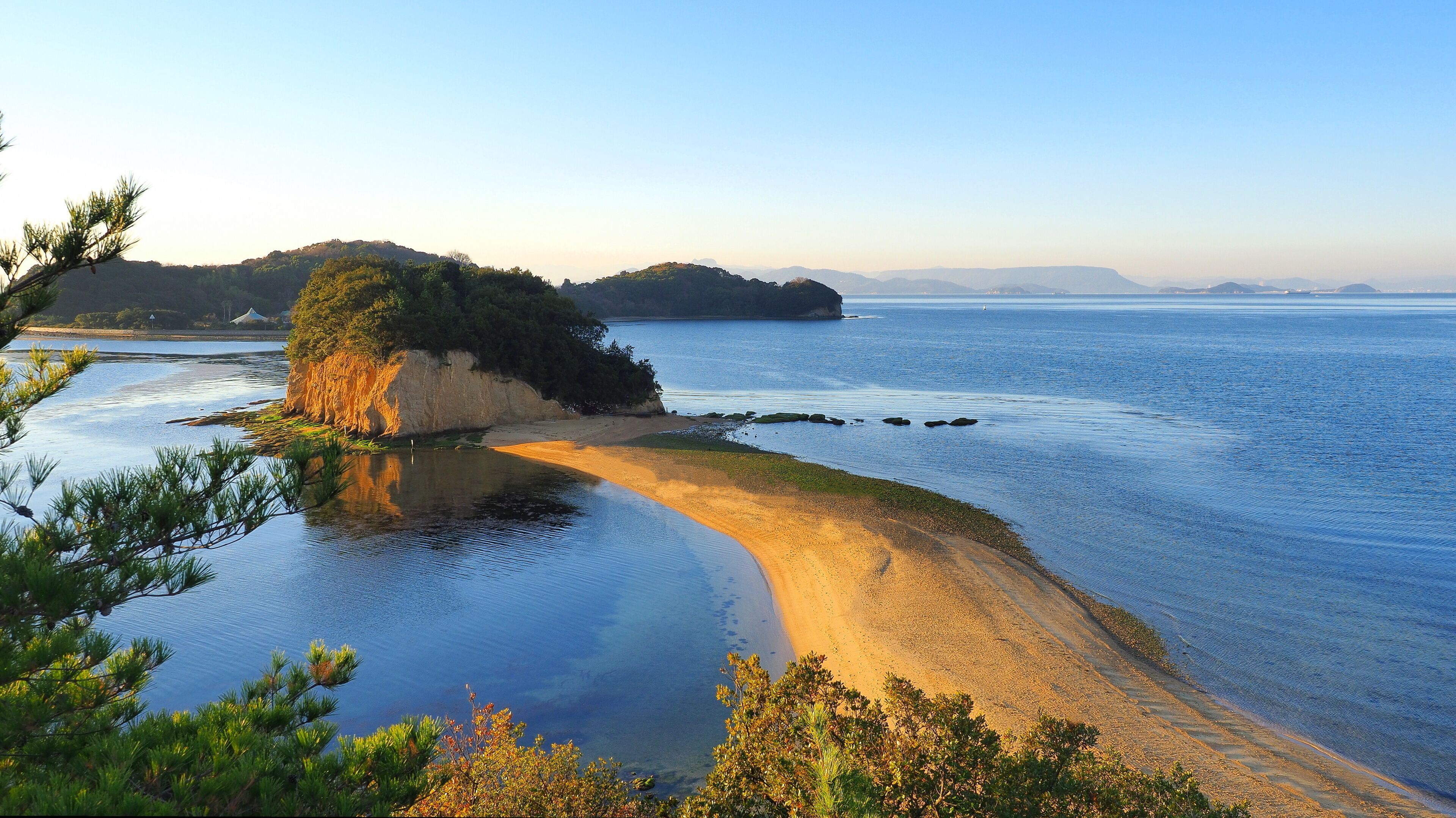 A coastal landscape with a sandy beach, calm waters, and a forested hill in the background.