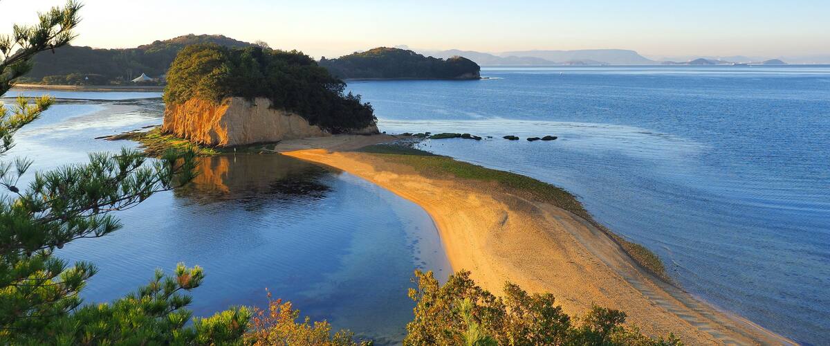 A coastal landscape with a sandy beach, calm waters, and a forested hill in the background.