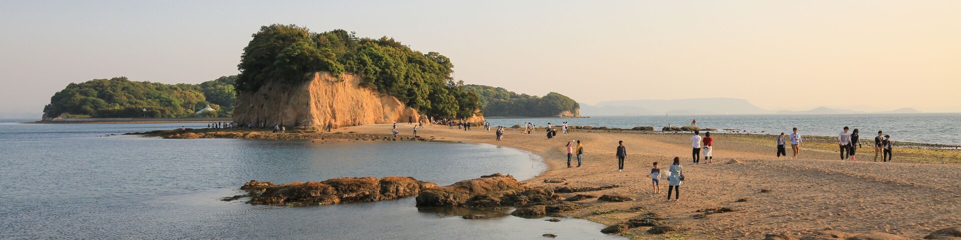 Una playa costera con costas rocosas, una pequeña isla con abundante vegetación y gente caminando por la orilla.