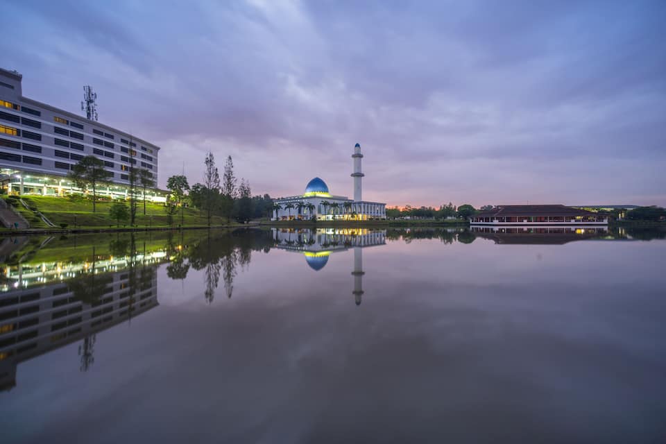 A mosque with a blue dome and a minaret, reflecting in a calm body of water, surrounded by modern buildings and greenery.
