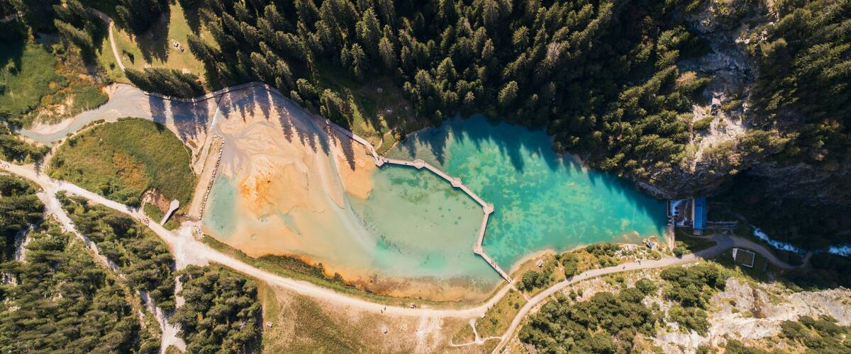 An aerial view of a coastal area with a clear turquoise bay, surrounded by dense forests and a rocky cliff.