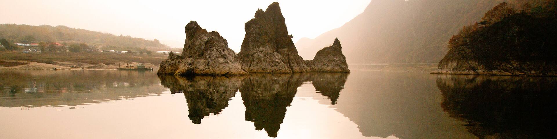 A calm lake with three rock formations in the center, surrounded by hills and a misty atmosphere.