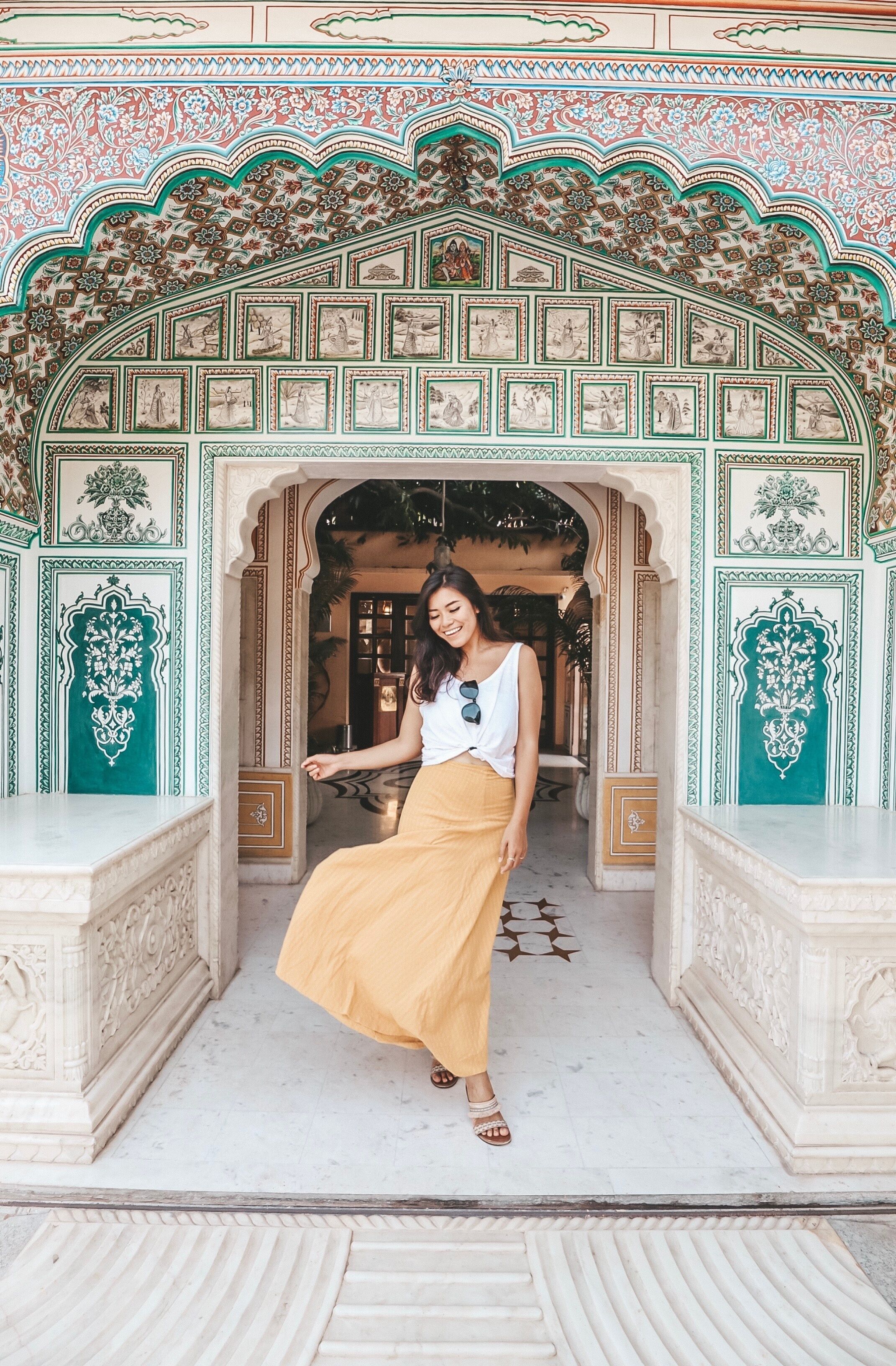 A woman in a white top and mustard skirt standing in front of an ornate archway with intricate designs and a tiled ceiling.