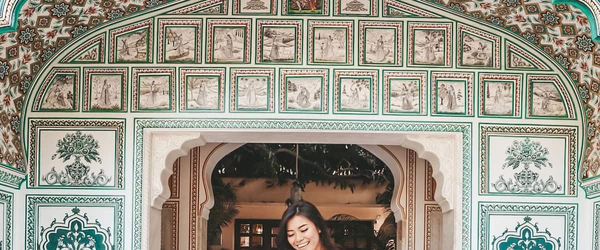 A woman in a white top and mustard skirt standing in front of an ornate archway with intricate designs and a tiled ceiling.