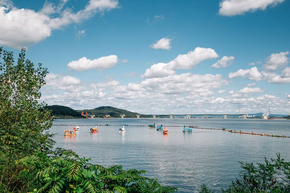 Um lago tranquilo com vários barcos, uma ponte ao longe e vegetação exuberante na margem.