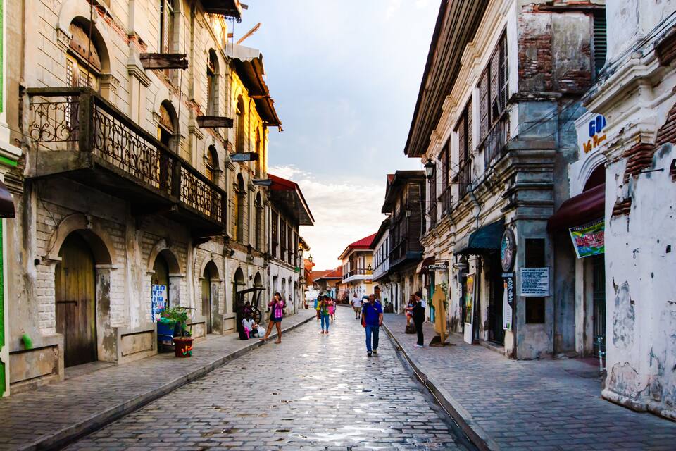 Uma rua de paralelepípedos ladeada por edifícios históricos, alguns com sacadas e placas.