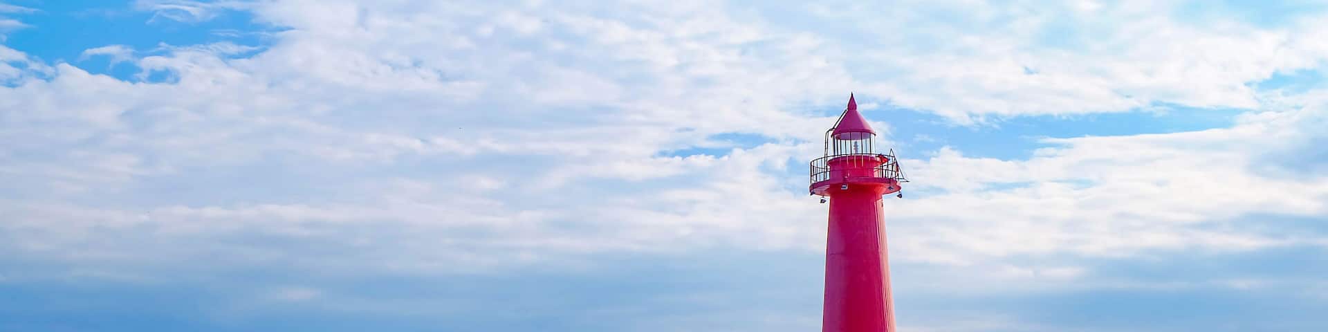 Un faro rojo sobre un promontorio rocoso, con aguas tranquilas y cielo despejado.