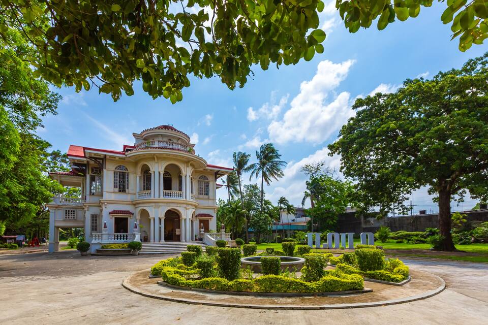 Un imposant bâtiment de style colonial, avec un toit rouge, entouré de jardins bien entretenus et de palmiers.