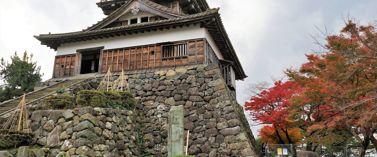 A traditional Japanese castle with a stone base and wooden upper structure, surrounded by autumn foliage.