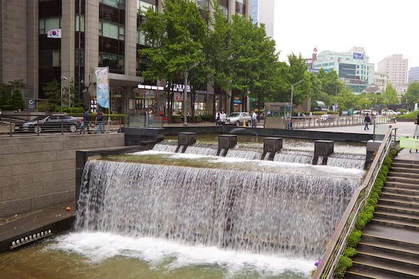 A multi-tiered waterfall in an urban setting with modern buildings and a colorful sculpture in the background.