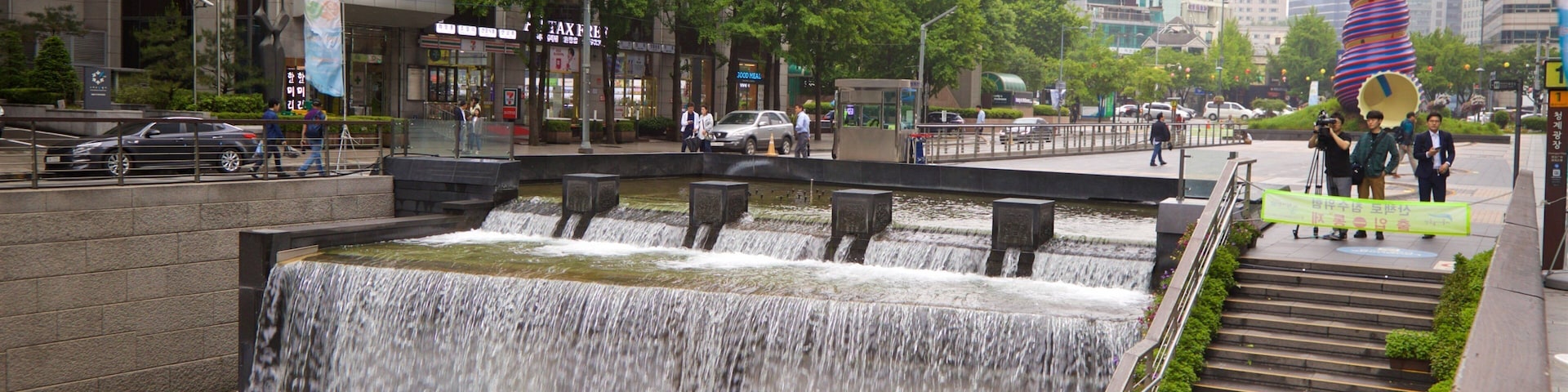 Uma cascata em vários níveis em ambiente urbano, com edifícios modernos e uma escultura colorida ao fundo.