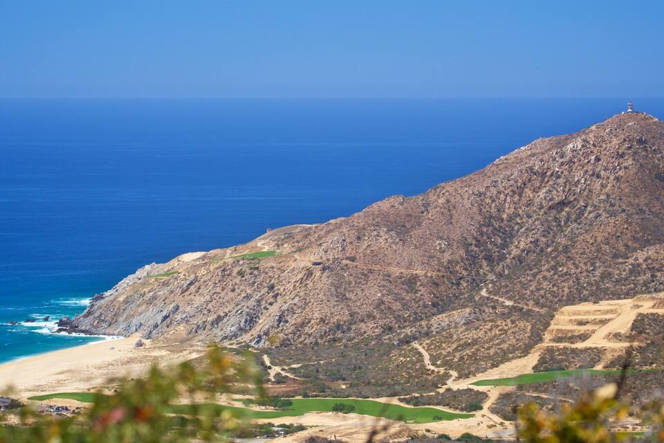 A rugged mountain with a lighthouse on top, overlooking a beach and a clear blue sea.