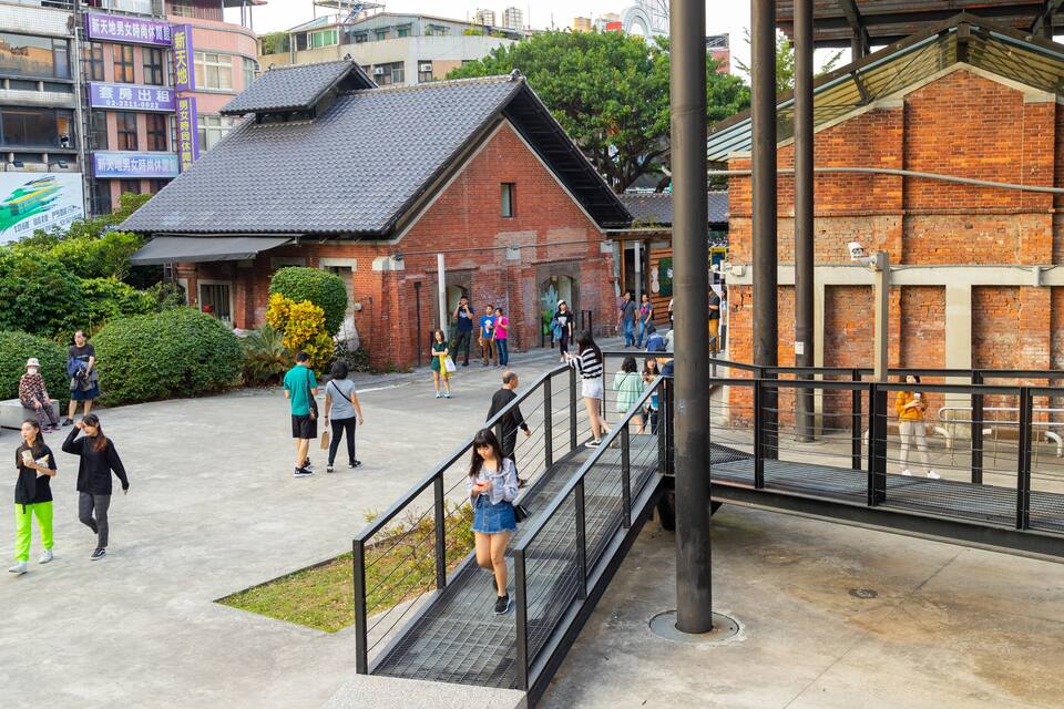 A public square with a brick building, a metal staircase, and people walking.