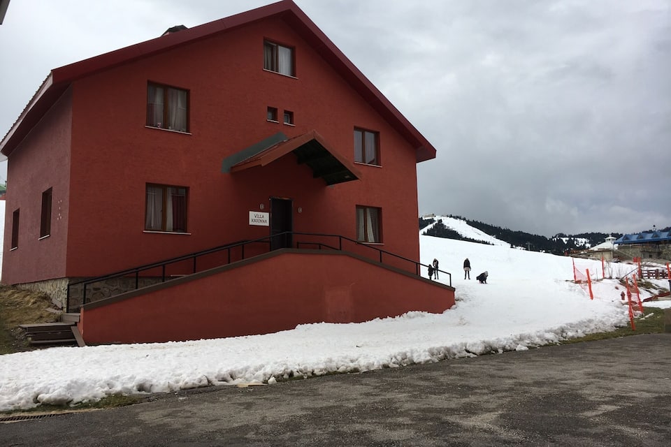 Une villa rouge avec un escalier et une enseigne indiquant « Villa Karinna », dans un paysage enneigé avec des montagnes en arrière-plan.