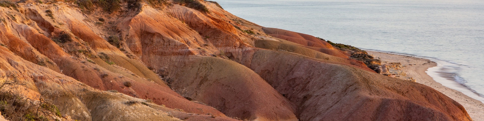 Paisagem costeira com falésias avermelhadas, uma praia de areia e um mar calmo.