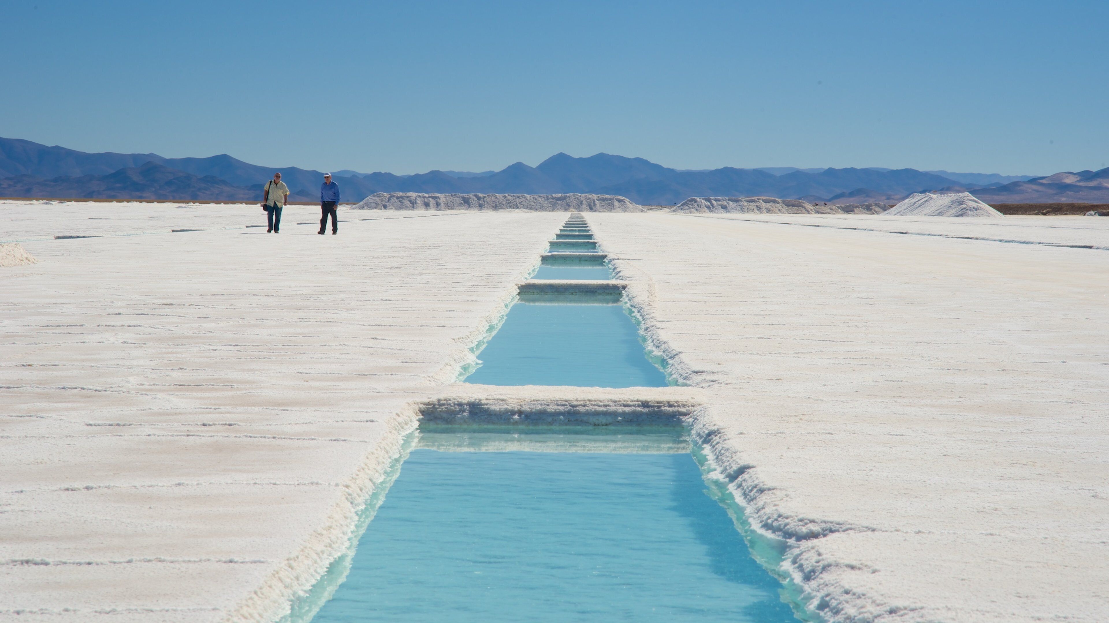 A salt flat with a blue water channel and mountains in the background.