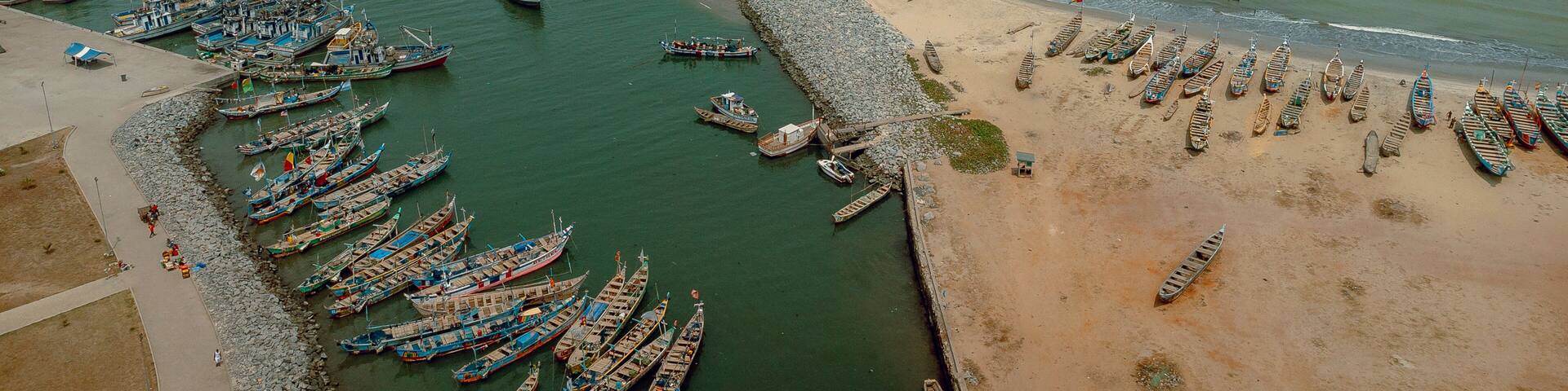 Aerial view of fishing boats clustered in the harbor contrasted against the long breakwater and sandy beach, Elmina, Central Region, Ghana.