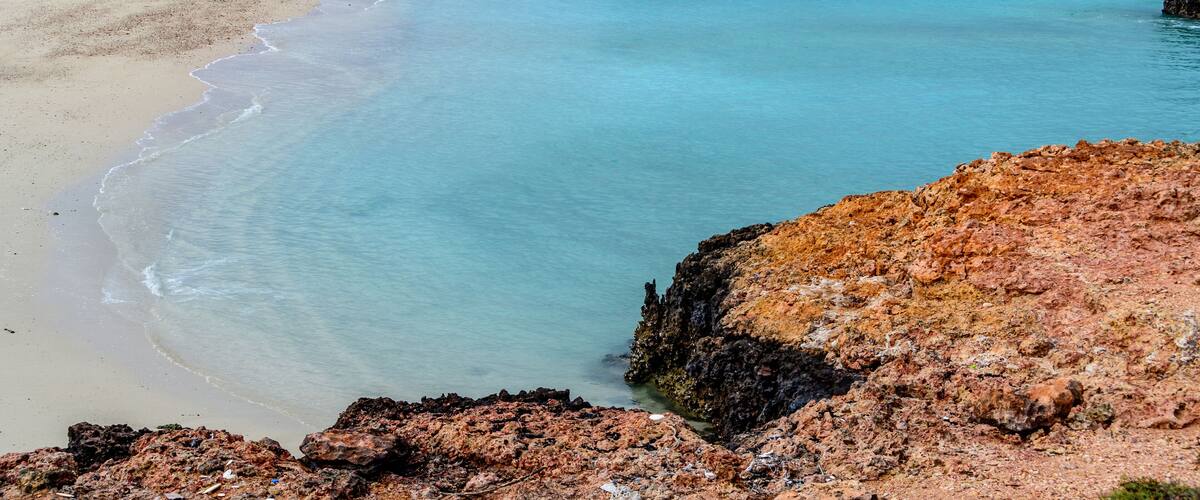 A coastal landscape with a sandy beach, clear turquoise waters, and rocky outcrops.