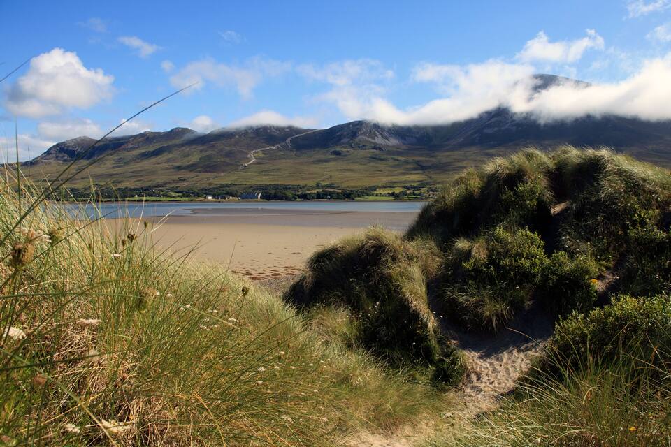 Un paysage côtier avec une plage de sable, des dunes herbeuses, un lac et des montagnes en arrière-plan.