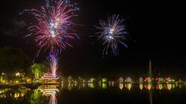 Fuegos artificiales sobre un lago tranquilo con edificios iluminados y una pagoda tradicional.