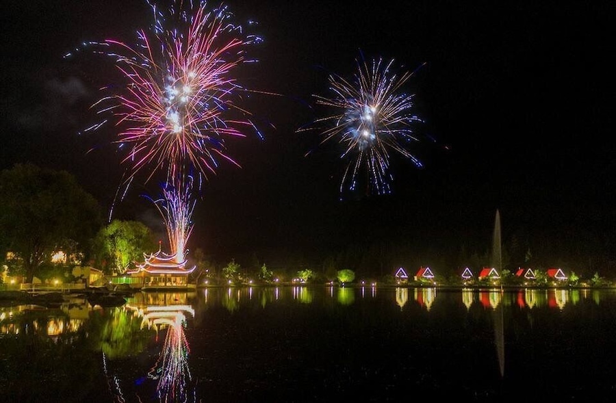 Fuegos artificiales sobre un lago tranquilo con edificios iluminados y una pagoda tradicional.