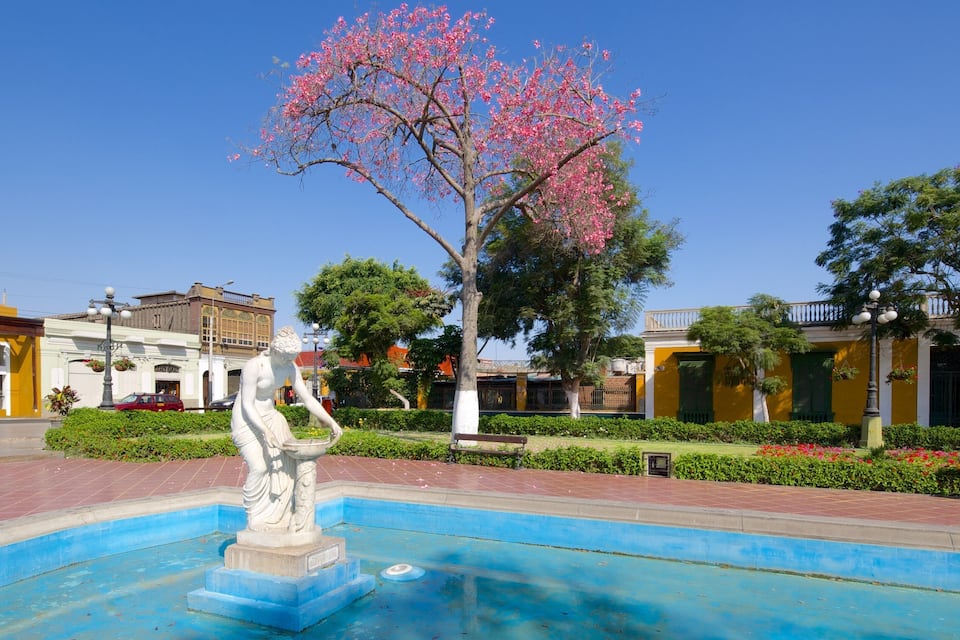 A fountain with a statue, surrounded by a blue pool, in a plaza with buildings and trees.