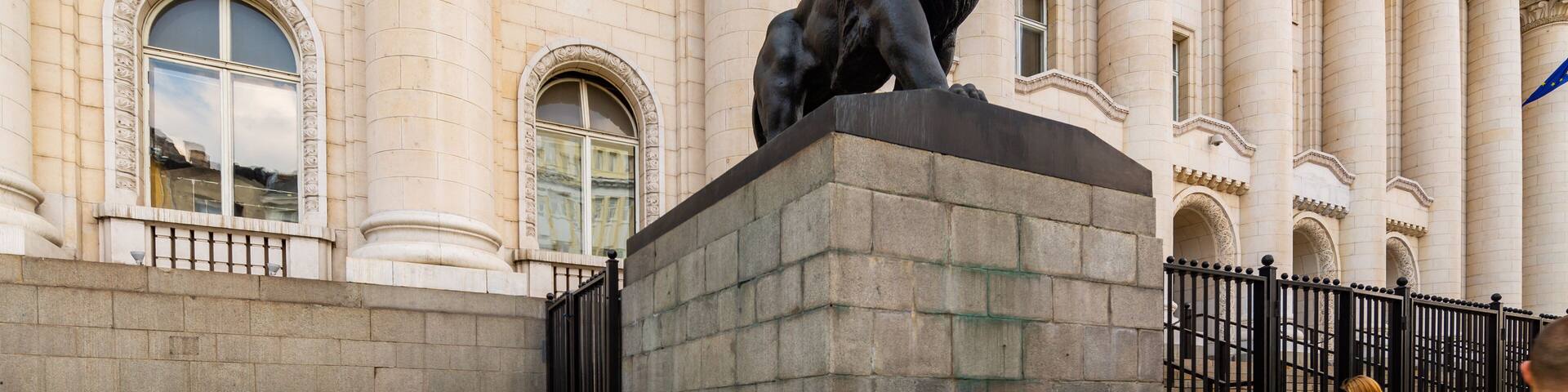 Una estatua de león sobre un pedestal frente a un edificio con columnas y la inscripción "ПАНАХА".
