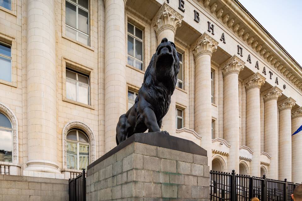 Una estatua de león sobre un pedestal frente a un edificio con columnas y la inscripción "ПАНАХА".