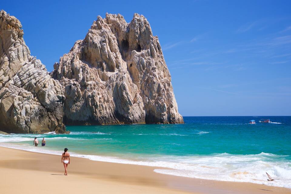 A beach with clear turquoise waters, rocky cliffs, and a few people walking along the shore.