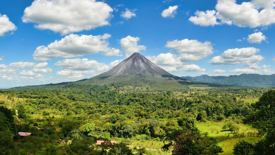A volcano surrounded by lush greenery and a clear blue sky with scattered clouds.