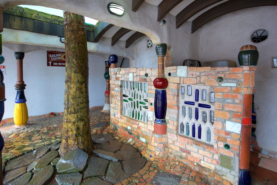 Un intérieur rustique agrémenté d’un arbre, de colonnes aux couleurs vives en forme de bouteille et d’un mur de briques percé de fenêtres en forme de bouteille.